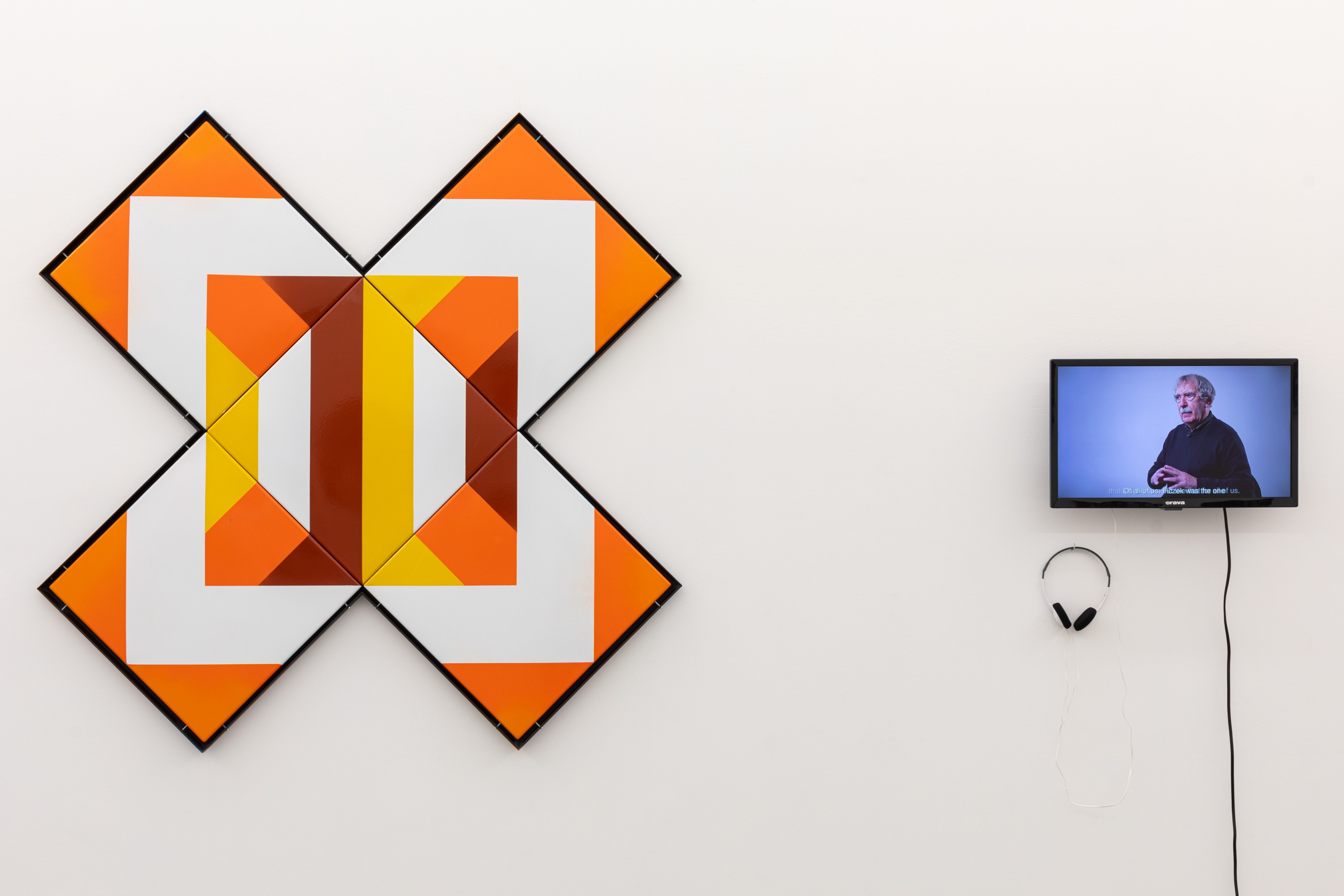 Pięć płytek ceramicznych z geometrycznym wzorem o ciepłych barwach, zawieszonych na ścianie w taki sposób, aby tworzyły znak "X". Obok wisi mały ekran, na którym wyświetlany jest film. Obok ekranu znajdują się czarne słuchawki.